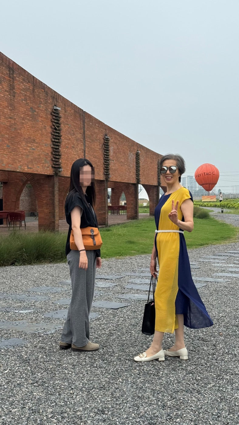 Privacy-protected travel reference photo showing a yellow-and-navy outfit, sunglasses, handbag, and peace sign