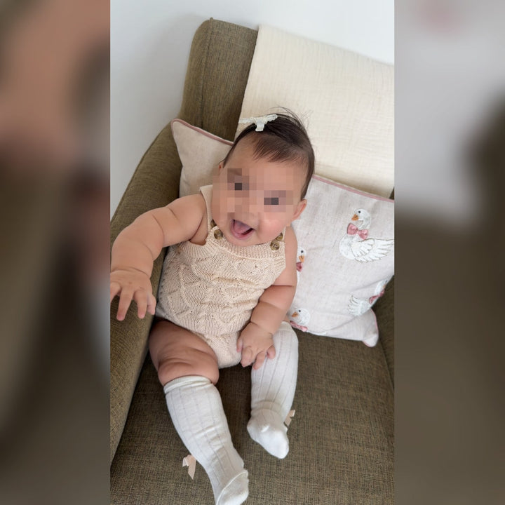 Privacy-protected reference photo of a seated baby reaching forward in a cream knitted romper and white bow socks