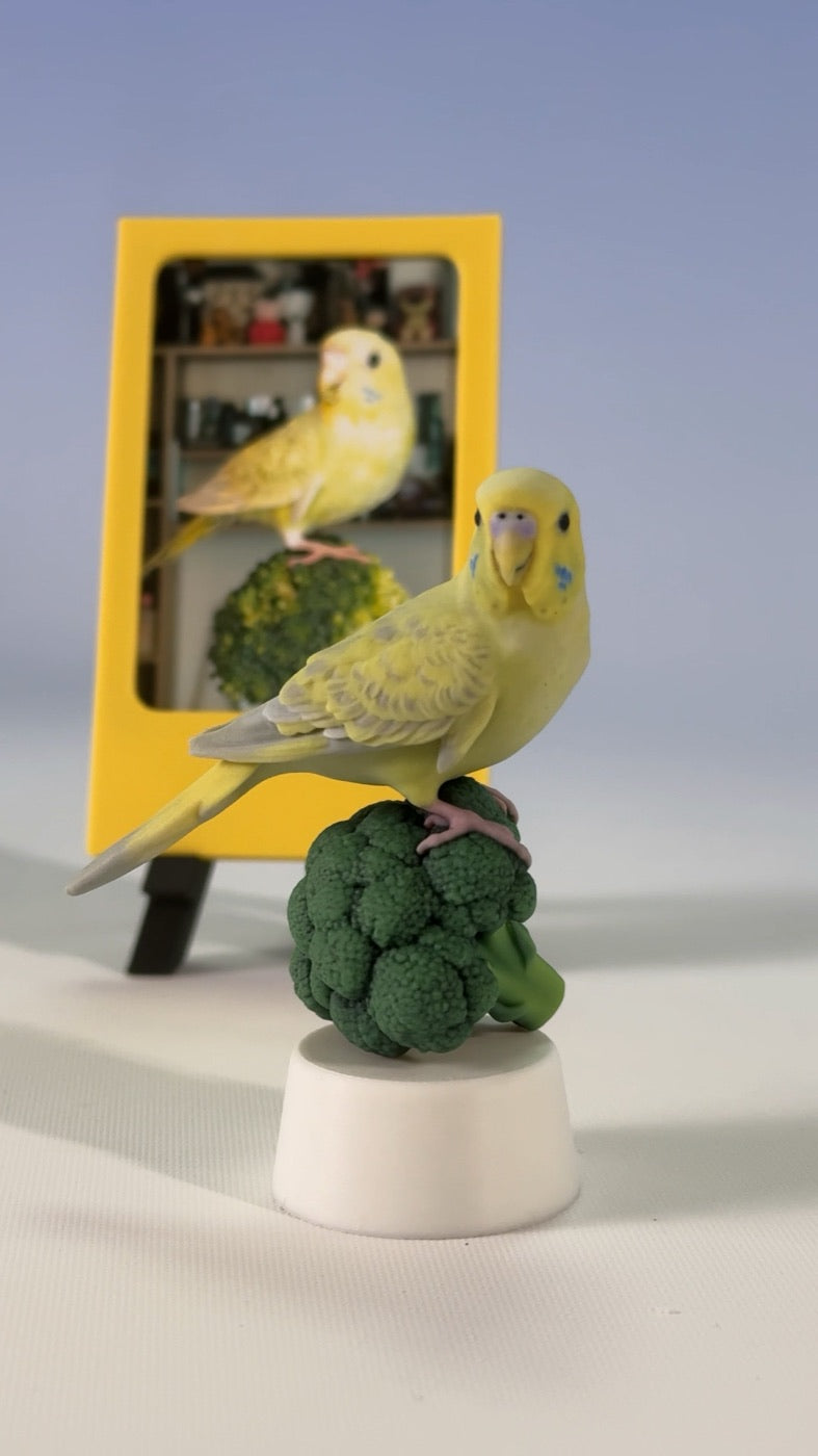 Finished custom yellow budgie figure on a broccoli perch beside a framed reference photo
